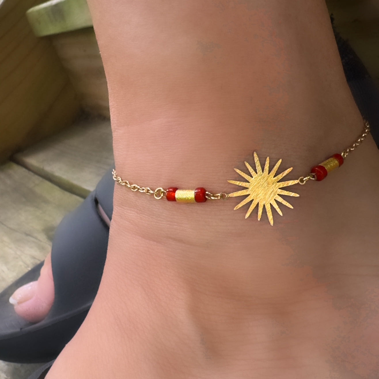 Gold anklet with sun design and red beads on a person's ankle.